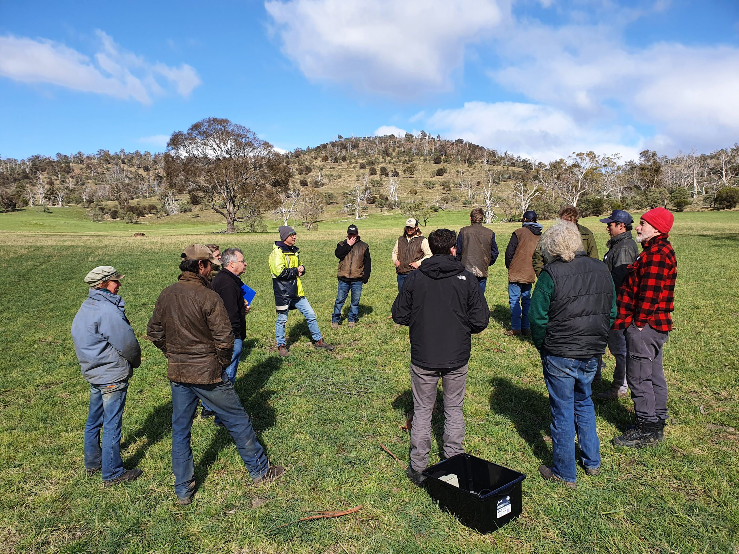 Group of people in a field standing in a circle and listening to a speaker